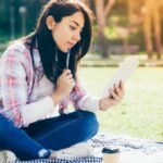 A student sitting cross-legged on a picnic blanket reads a letter on her smartphone