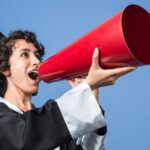 Stock photo of female student in graduation gown speaking into an old-timey megaphone